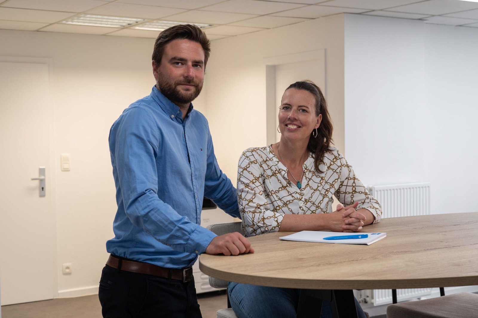 Een man staat naast een vrouw aan een ronde tafel met een blocnote en pen, beide met het gezicht naar de camera in een modern kantoor.