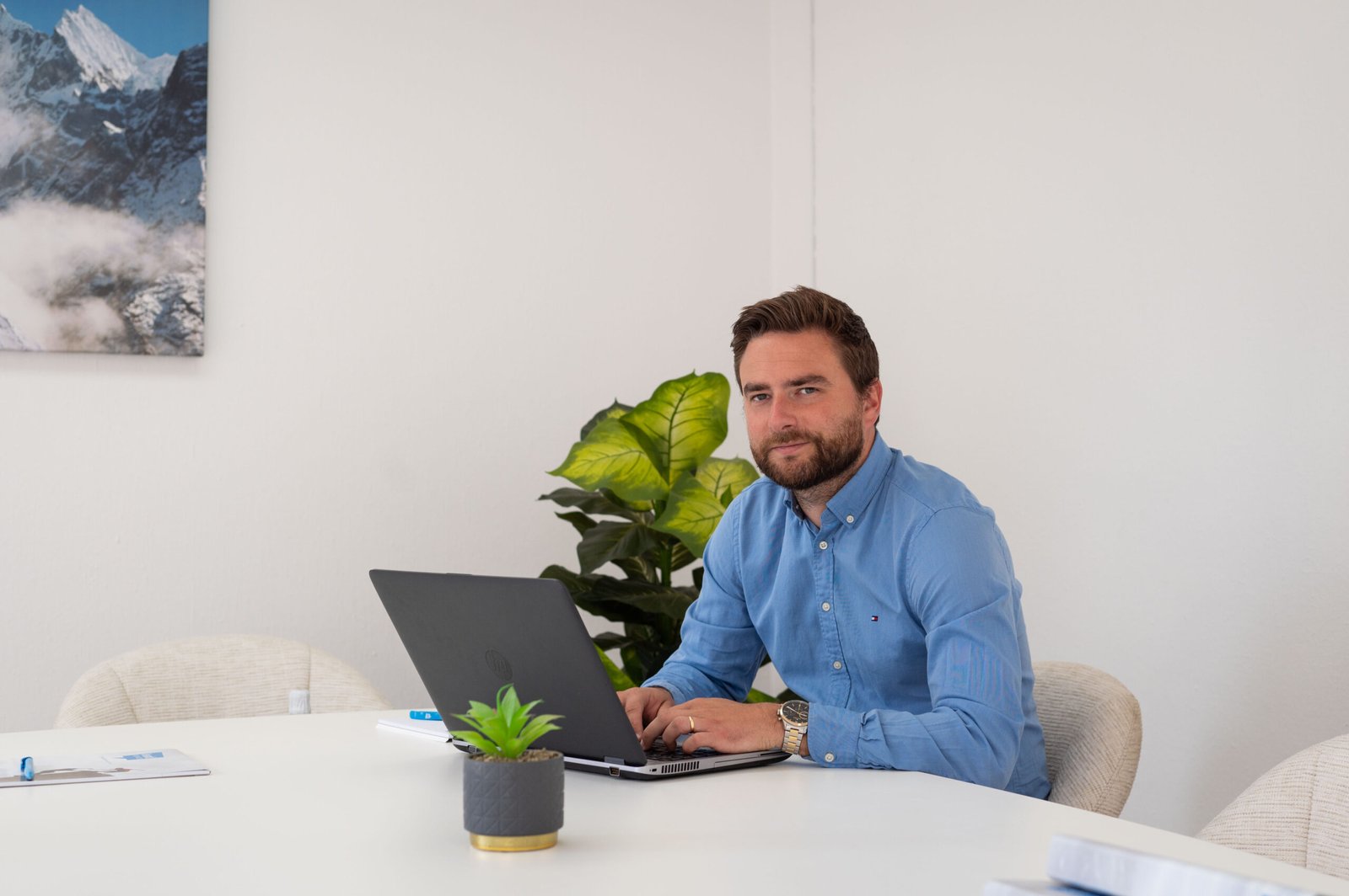 Een man in een blauw shirt zit aan een witte tafel te werken op een laptop, met een potplant en muurkunst op de achtergrond.